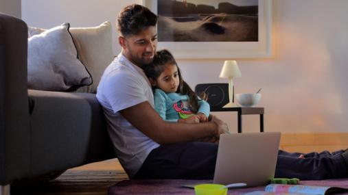 Father sat on the floor with his daughter on his knee, happily looking at a computer screen.